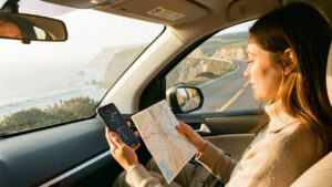 Une voyageuse assise dans une voiture sur une route côtière compare un itinéraire sur son téléphone avec une carte papier, éclairée par la lumière douce du matin.